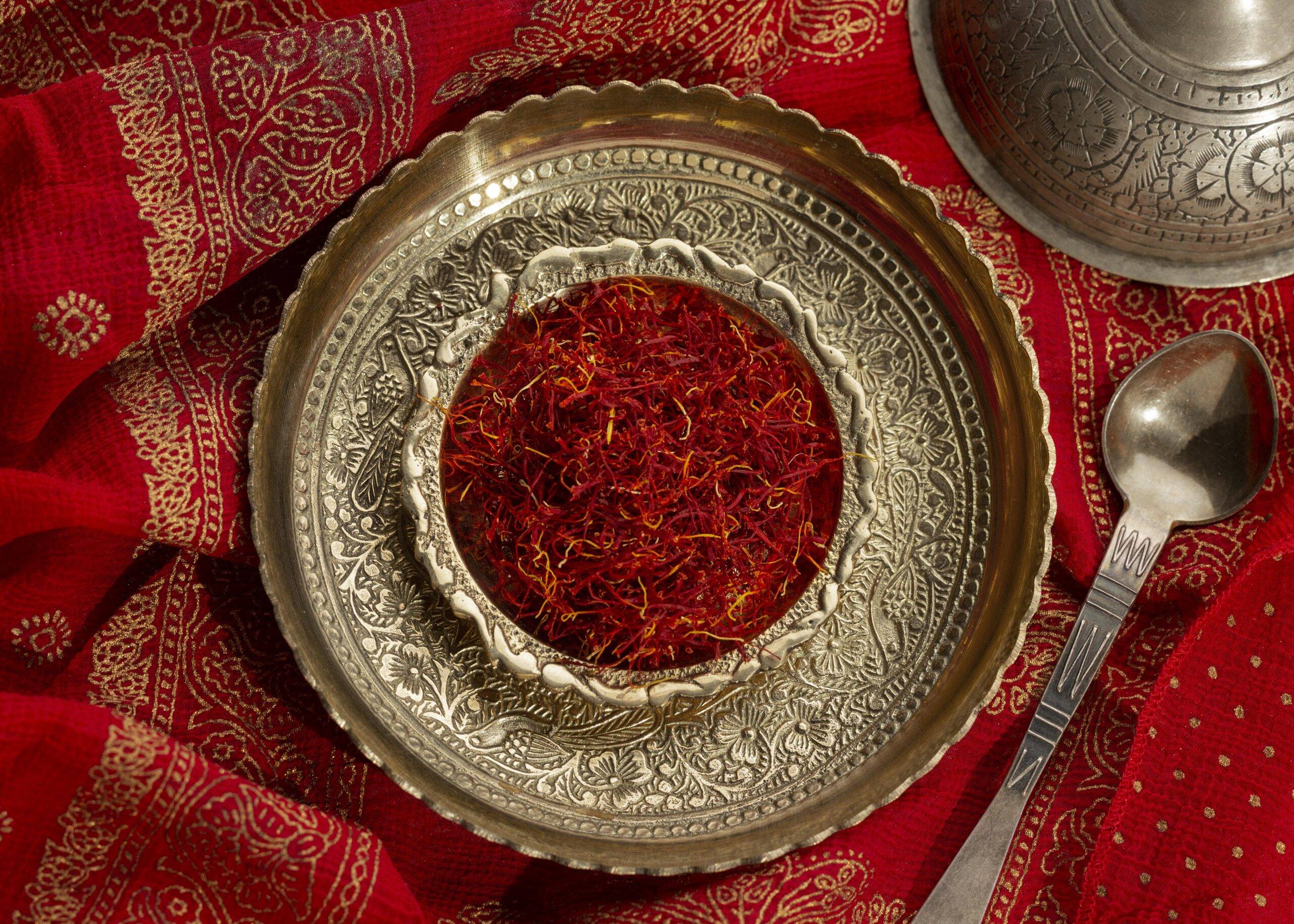 A decorative brass plate filled with vibrant red saffron threads, placed on an ornate red and gold patterned fabric, accompanied by an intricately designed metal spoon.